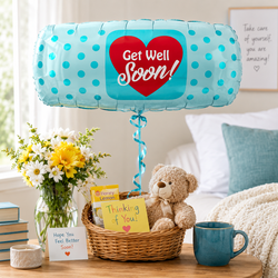 A round foil balloon with turquoise blue polka dots and a large red heart in the center that reads 'Get Well Soon!' in white script, floating above a bedside table. The table holds a wicker basket with a teddy bear, a yellow card, a mug, flowers, and a small sign, set in a softly lit bedroom.