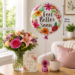 A round foil balloon with a floral design and the words 'Feel Better Soon' stands on a table next to a vase of flowers, a card, and a mug. The scene is set in a bright living room with a sofa, and a measurement overlay notes the balloon is 18 inches (45 cm) tall.