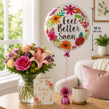 A round foil balloon with a floral design and the words 'Feel Better Soon' stands on a table next to a vase of flowers, a card, and a mug. The scene is set in a bright living room with a sofa, and a measurement overlay notes the balloon is 18 inches (45 cm) tall.