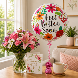 A round foil balloon with colorful floral illustrations and the words 'Feel Better Soon' stands on a table beside a vase of pink flowers, a greeting card that says 'Thinking of You,' and a pink tinsel ornament in a cozy living room setting.