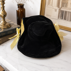 A black velvet witch-style hat with gold trim and gold ribbon bows, displayed on a white marble surface with books and a brass object in the background.