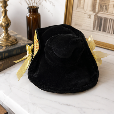 A black velvet witch-style hat with gold trim and gold ribbon bows, displayed on a white marble surface with books and a brass object in the background.