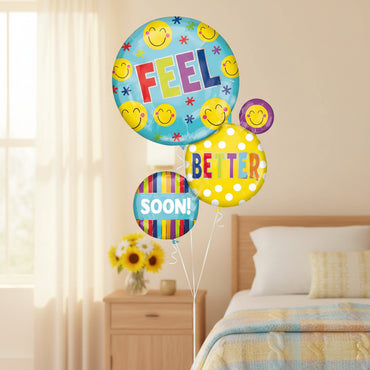 A set of round party balloons with a happy birthday theme, featuring smiley faces, the word 'FEEL' on a light blue balloon, 'BETTER' on a yellow polka-dot balloon, and 'SOON!' on a striped balloon.
