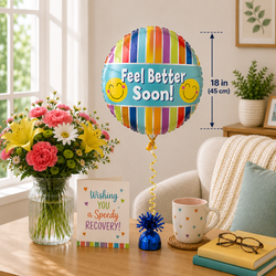 A colorful striped round foil balloon with the words 'Feel Better Soon!' and smiley faces, shown in a styled living room setting next to a bouquet of flowers, a greeting card, a mug, and books. A dimension marker on the right indicates the balloon is 18 in (45 cm) tall.
