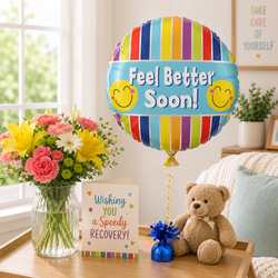 A colorful foil balloon with vertical rainbow stripes and the text 'Feel Better Soon!' with two yellow smiley faces, displayed on a wooden table next to a bouquet of flowers, a teddy bear, and a recovery card in a bright living room setting.