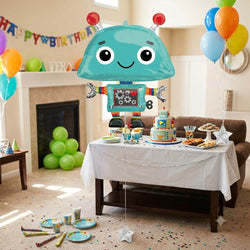 A large teal robot-shaped balloon with red antenna tips is floating above a decorated children's birthday party table. The room has a fireplace, balloons in multiple colors, a banner reading Happy Birthday, and party supplies on the floor and table.