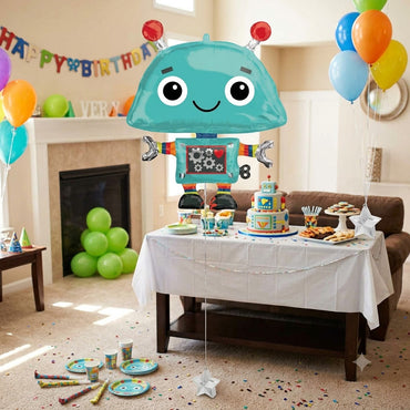A large teal robot-shaped balloon with red antenna tips is floating above a decorated children's birthday party table. The room has a fireplace, balloons in multiple colors, a banner reading Happy Birthday, and party supplies on the floor and table.