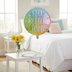 A round foil balloon with a pastel rainbow gradient background and the words 'FEEL BETTER SOON' in large white script with a light blue outline. Small gold confetti dots are scattered across the balloon, and the balloon is shown on a plain white background.