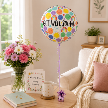 A round foil balloon with a colorful smiley-face and rainbow pattern and the words 'GET WELL SOON!' floating above a wooden table in a cozy living room setting. The table is styled with a bouquet of pink and white flowers, a mug, a small card, and a decorative pom-pom, next to a beige armchair with a cream knit throw.
