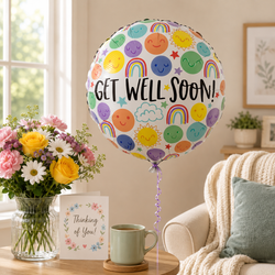 A round foil balloon with a white background, colorful smiling suns, rainbows, clouds, and stars, printed with the words 'GET WELL SOON!' in black. The balloon is shown in a cozy living room setting next to a vase of flowers, a card that says 'Thinking of You!', and a mug.