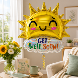 A gold foil balloon shaped like a smiling sun with the words 'GET WELL SOON!' on a white scalloped base, displayed in a bright living room setting next to a vase of sunflowers and a mug.