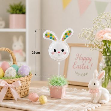 A white bunny head balloon decoration on a stick, displayed in a pink pot with artificial grass on a table. The image includes a size measurement overlay indicating 23cm, surrounded by Easter-themed props like a basket of decorated eggs, a ceramic bunny, and a 'Happy Easter' sign.