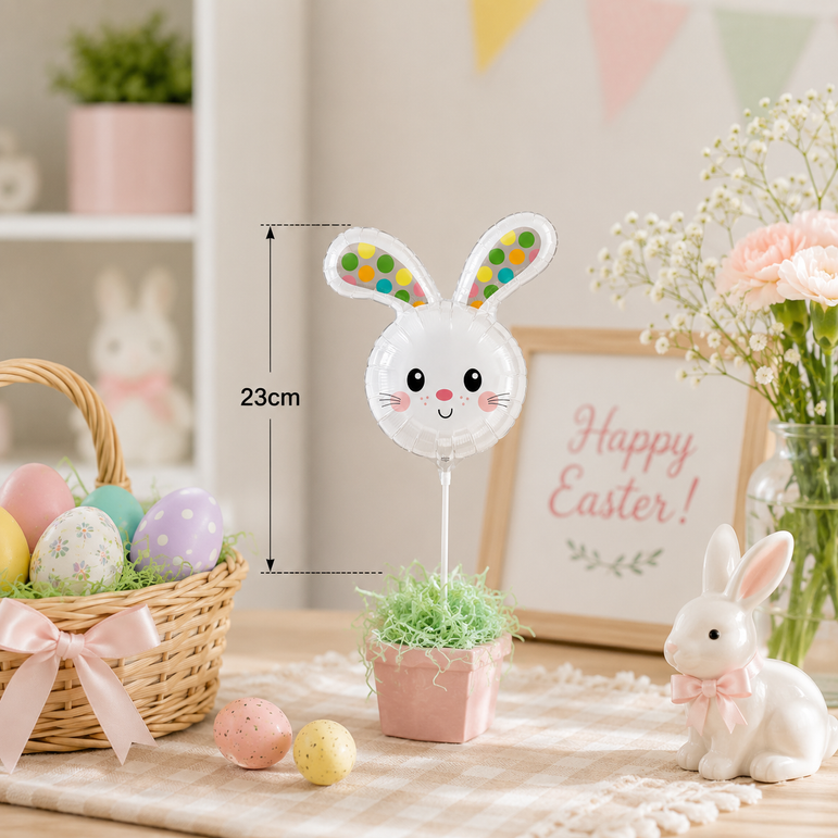 A white bunny head balloon decoration on a stick, displayed in a pink pot with artificial grass on a table. The image includes a size measurement overlay indicating 23cm, surrounded by Easter-themed props like a basket of decorated eggs, a ceramic bunny, and a 'Happy Easter' sign.