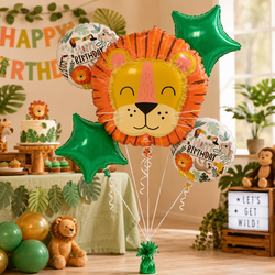 A lion-themed birthday balloon bouquet with a large orange and yellow lion face balloon, green star balloons, and smaller safari-themed round balloons, displayed in a decorated children's party setting with a cake, plants, and stuffed animals.