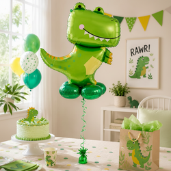 A large green dinosaur-shaped foil balloon floating above a decorated party table. The scene includes a green and white cake, matching party cups, a paper bag, bunting, and a framed dinosaur print in a bright indoor party setting.