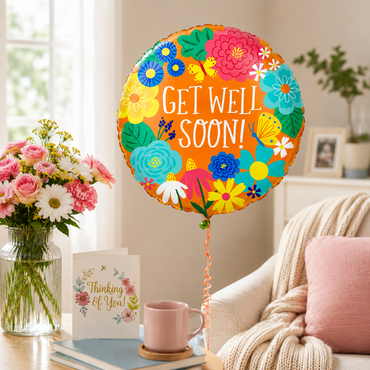 A round foil balloon with a colorful floral border and the words 'GET WELL SOON!' floating in a bright living room setting next to a vase of flowers, a card, and a pink mug.
