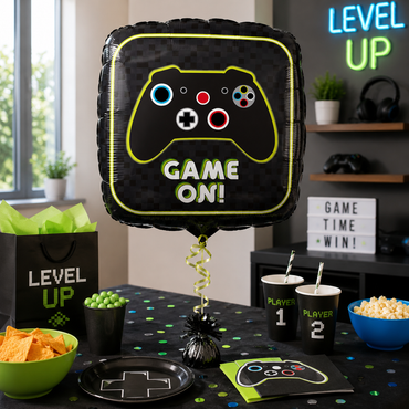 A black game controller themed party balloon reading 'GAME ON!' stands on a table in a decorated party setting with matching cups, plates, popcorn, chips, and neon-style decor in the background.