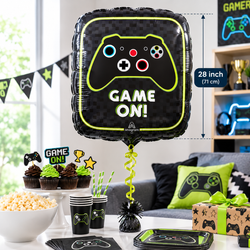 A black inflatable foil balloon with a neon green video game controller graphic and the words 'GAME ON!' is shown in a party setup. The image includes dimension callouts indicating the balloon is 28 inch (71 cm), with matching game-themed party supplies like cups, plates, cupcakes, napkins, and pillows on a couch in the background.