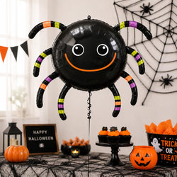 A large black spider-shaped foil balloon with a smiling face and striped legs stands on a decorated Halloween table. The scene includes pumpkins, cupcakes, orange tissue paper, a lantern, a spider web wall hanging, and a 'Happy Halloween' sign.