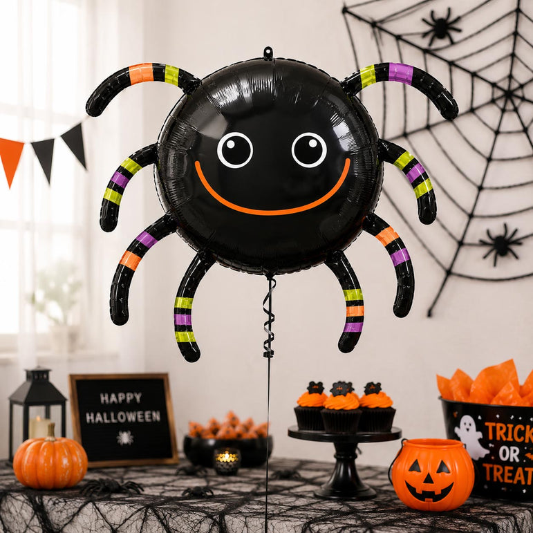 A large black spider-shaped foil balloon with a smiling face and striped legs stands on a decorated Halloween table. The scene includes pumpkins, cupcakes, orange tissue paper, a lantern, a spider web wall hanging, and a 'Happy Halloween' sign.