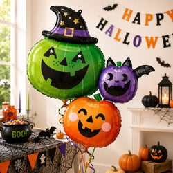 Three Halloween-themed foil balloons shaped like a green witch pumpkin, an orange jack-o'-lantern, and a purple bat-faced pumpkin, displayed in a decorated living room with a 'HAPPY HALLOWEEN' banner and pumpkins.
