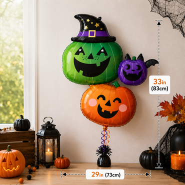 Three Halloween-themed foil balloons shaped like pumpkins with jack-o'-lantern faces are displayed on a tabletop against a beige wall. The balloons include a green pumpkin wearing a purple witch hat, an orange pumpkin with pink cheeks, and a smaller purple pumpkin.