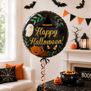 A round black foil balloon with a Halloween design, including the words 'Happy Halloween!' in gold, a white ghost, pumpkins, spiderwebs, and orange and black ribbons, standing in a decorated living room.