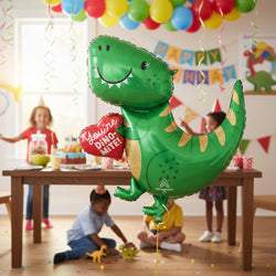 A green dinosaur-shaped foil balloon holding a red heart balloon that says 'You're Dino-Mite!' in white text. The balloon has gold accents on its back and belly, black facial features, and the Anagram logo near the bottom.