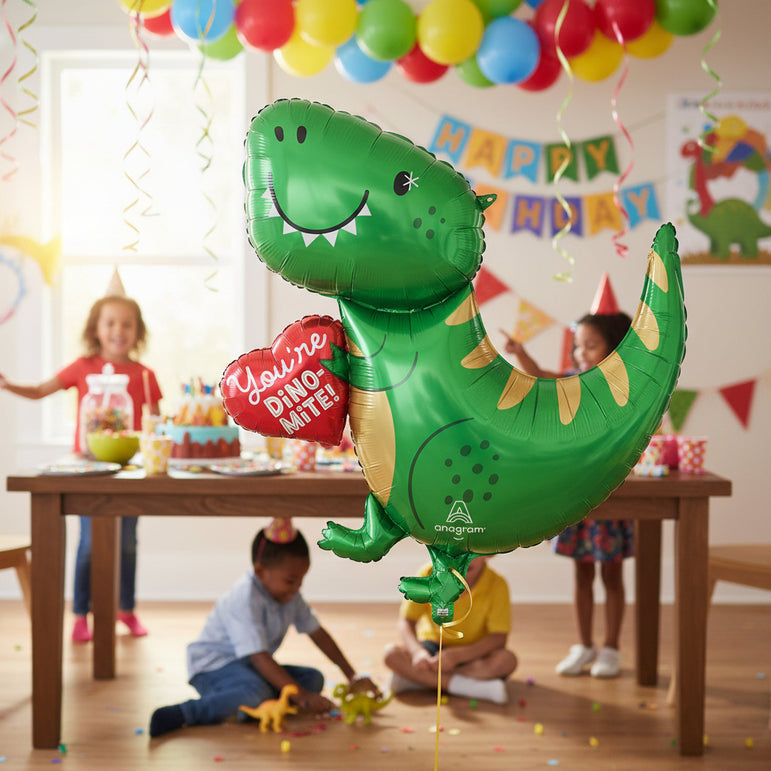 A green dinosaur-shaped foil balloon holding a red heart balloon that says 'You're Dino-Mite!' in white text. The balloon has gold accents on its back and belly, black facial features, and the Anagram logo near the bottom.